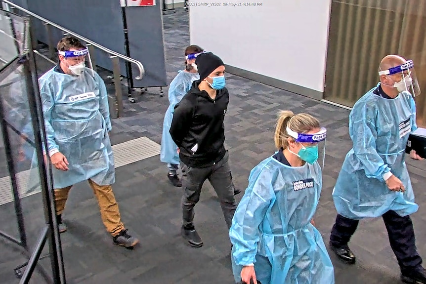 A young man in dark clothing and a surgical face mask, is escorted through an airport by four people in protective equipment.