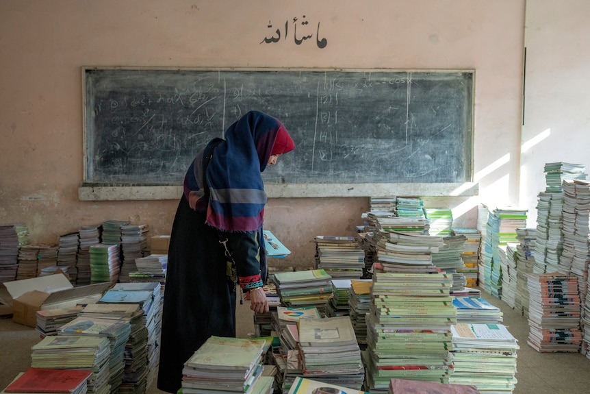 A woman wearing a head scarf packs up piles of books inside a classroom, a blackboard behind her