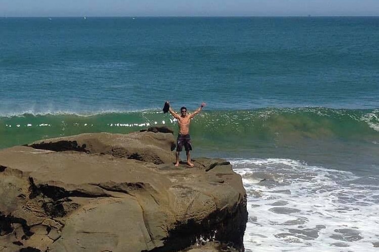 A shirtless man stands on a rock ledge with his arms raised as a wave crests behind him.