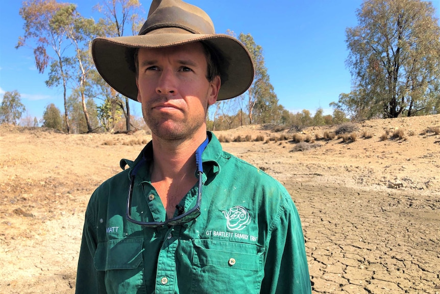 Matt Bartlett is wearing an akubra hat and standing on his dry dam bed, staring into the blue sky