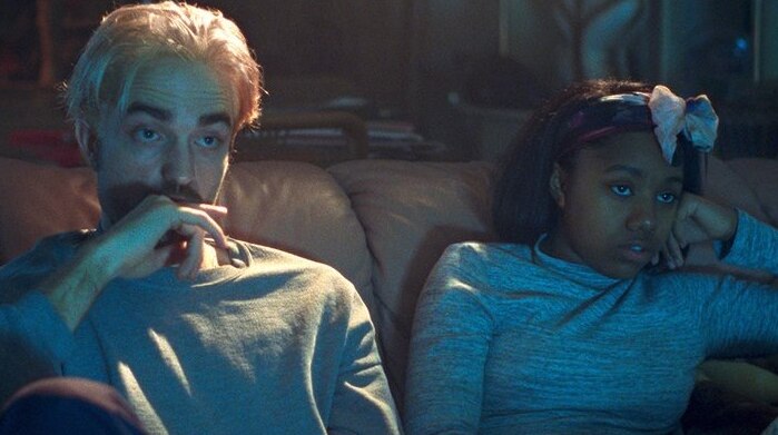 Still colour image from 2017 film Good Time, Robert Pattinson and Taliah Webster are seated on a couch in a dimly lit room.