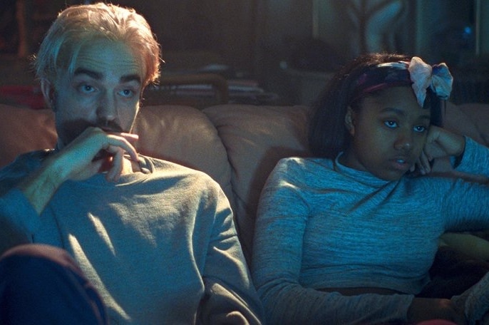 Still colour image from 2017 film Good Time, Robert Pattinson and Taliah Webster are seated on a couch in a dimly lit room.