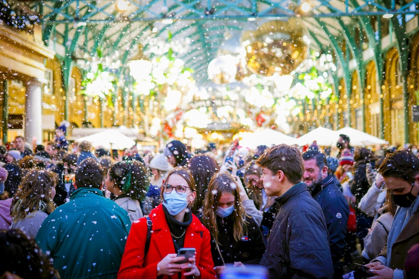 A woman in a red coat and blue face mask stands in a snowy, crowded market