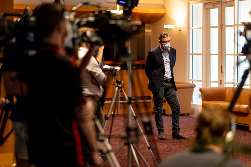 A man wearing a suit and a facemask waits to speak at a press conference. He looks serious.