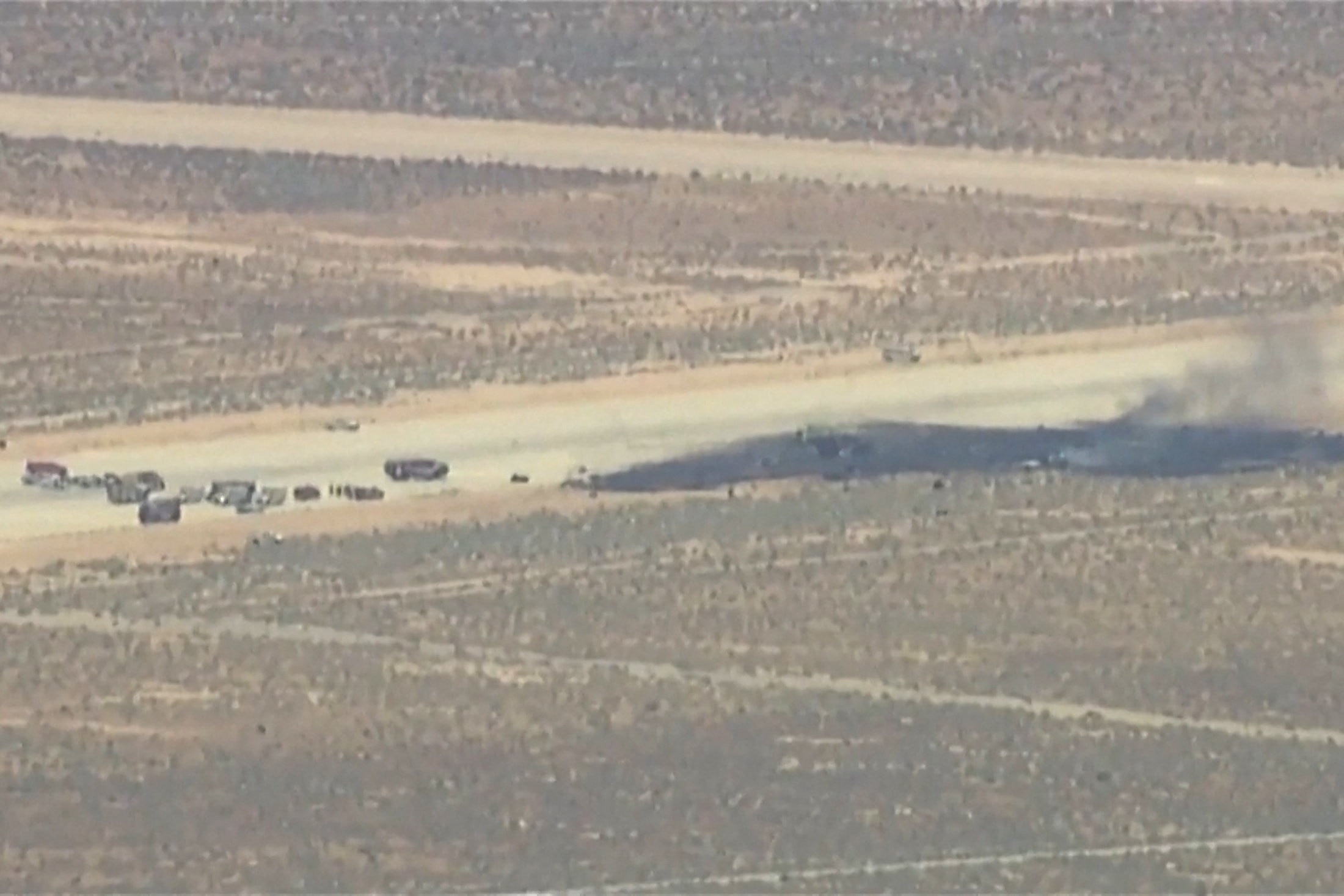 A long black burn mark on an air base runway smouldering with black smoke near parked vehicles.