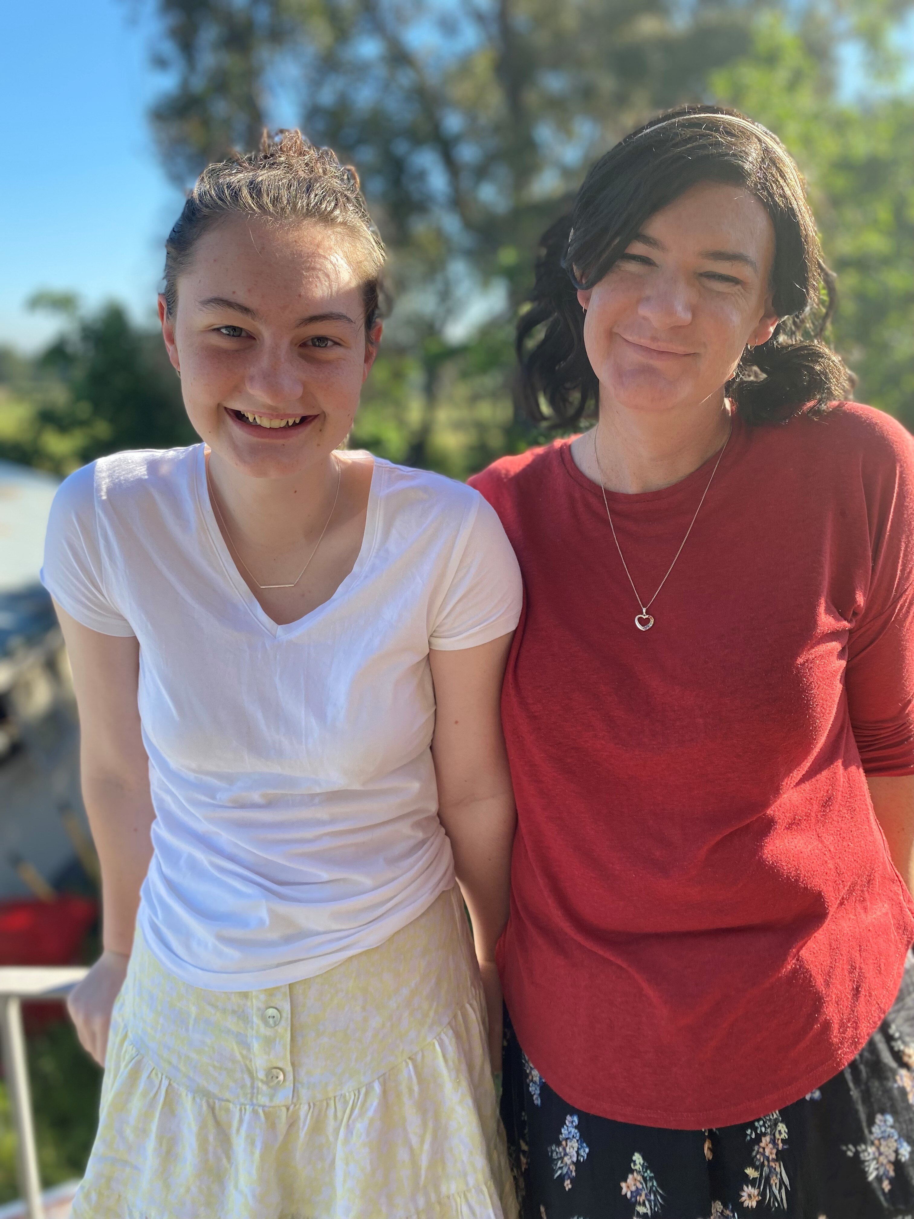 Mackenzie and Claire smile on a balcony in front of bushland. Both have medium-length hair, tied back.