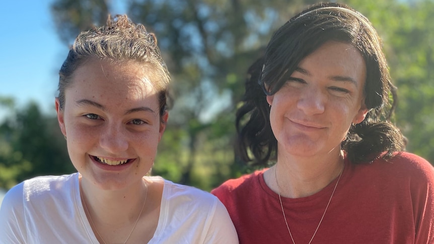Mackenzie and Claire smile on a balcony in front of bushland. Both have medium-length hair, tied back.