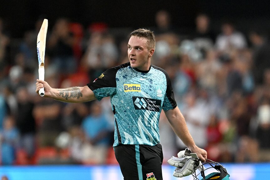 A male cricketer, wearing teal and black, raises his bat to thank the crowd as he leaves the field.