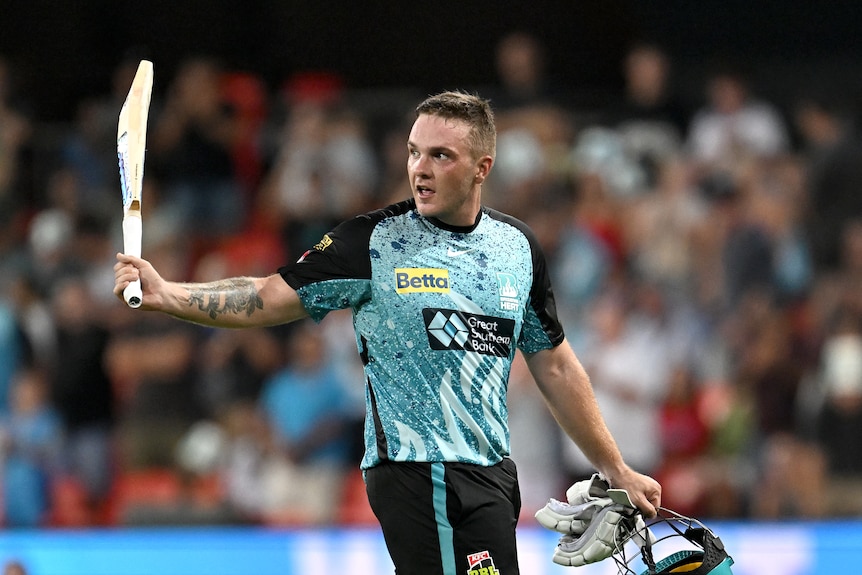 A male cricketer, wearing teal and black, raises his bat to thank the crowd as he leaves the field.