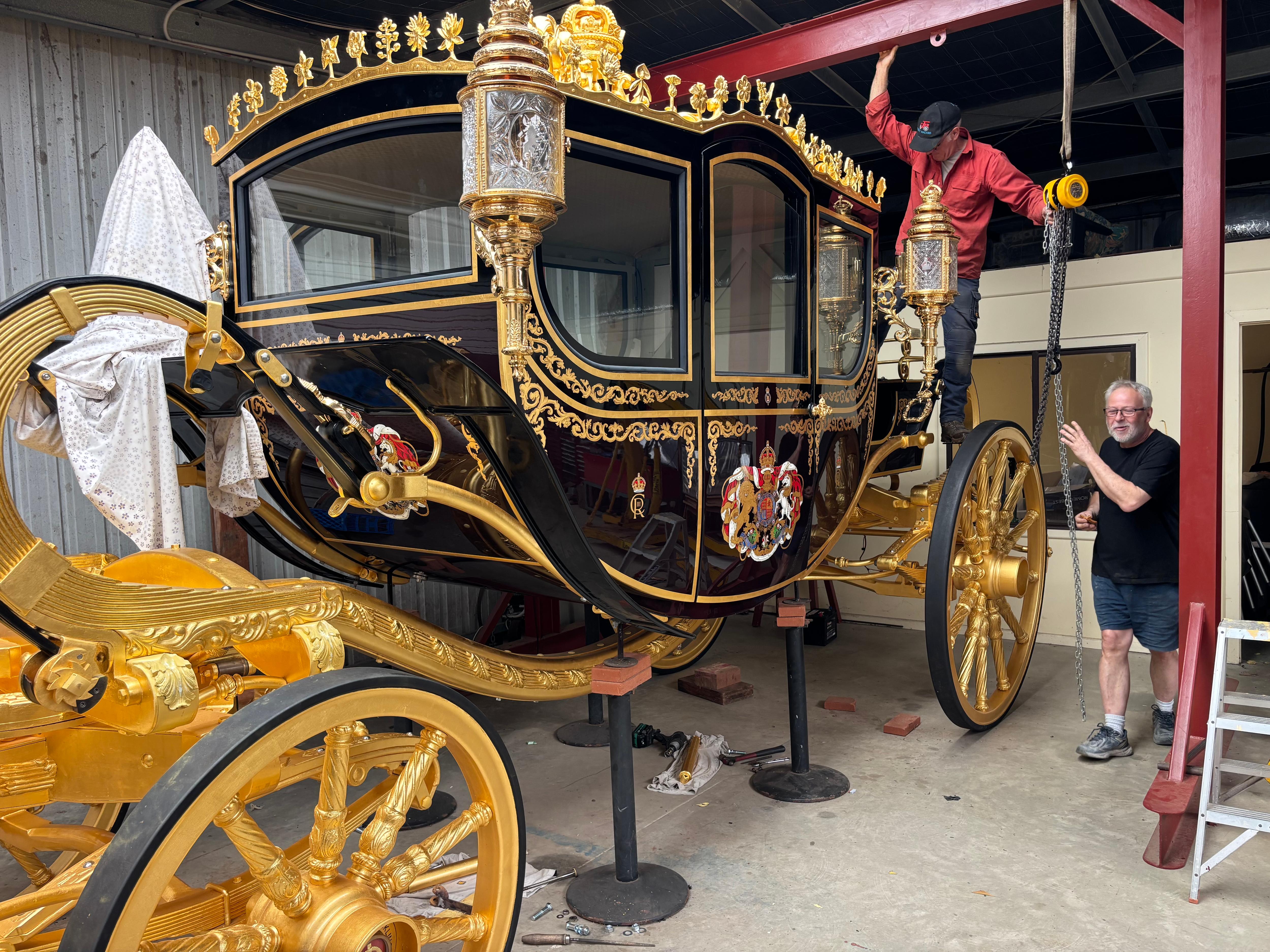 Two men work on a grand, gold and black royal coach in a shed.