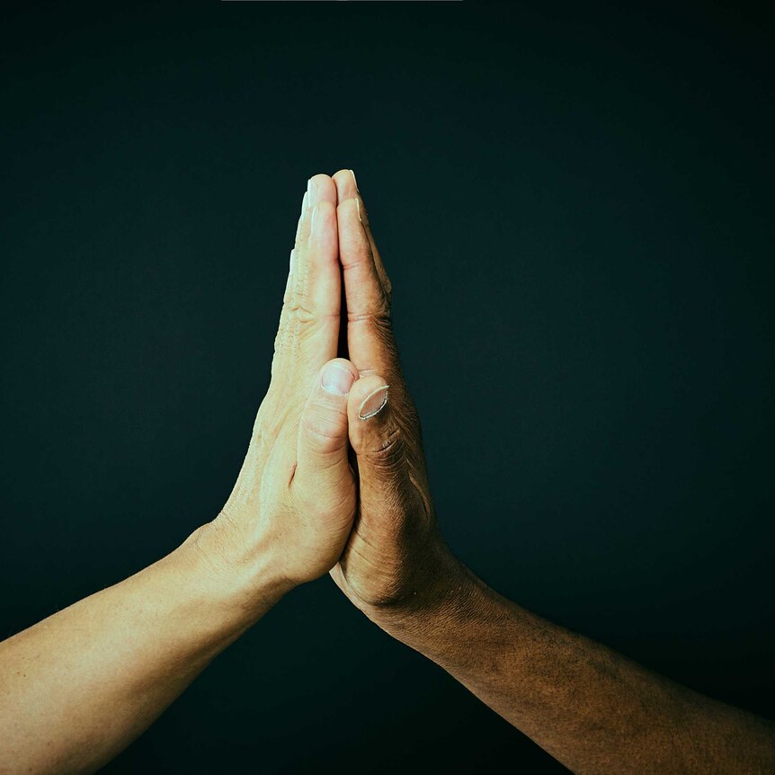 Two different men's hands come together to form a prayer symbol against a black background.