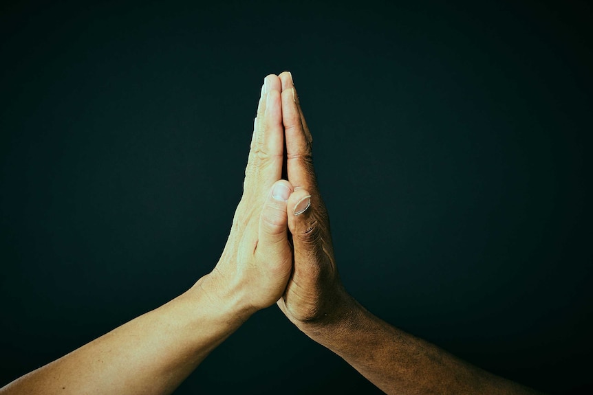 Two different men's hands come together to form a prayer symbol against a black background.