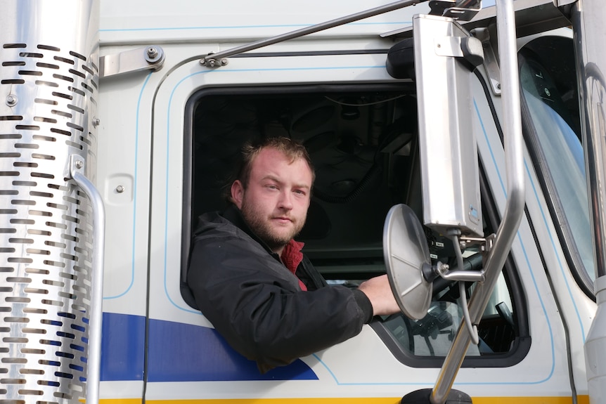 A driver with dark blonde hair and black pullover sits in his truck with his shoulder and arm out the window