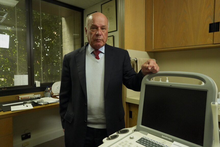 A balding man wearing a tie, knitted jumper and blazer stands by a medical machine.