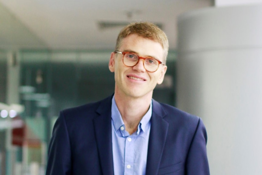 Michael Palmer, who has light brown hair, poses for a photo in a navy blazer, blue shirt and tortoiseshell spectacles