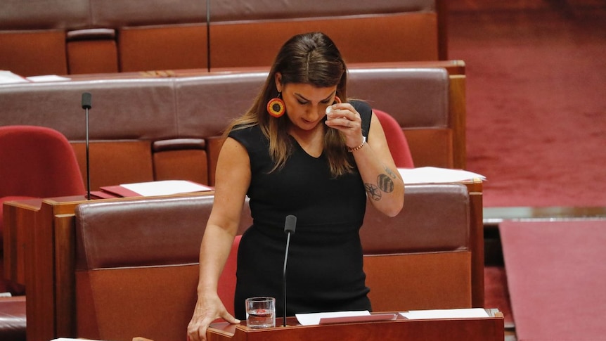 A dark haired woman in a black dress holds a tissue to her eye as she speaks in the Senate.