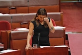 A dark haired woman in a black dress holds a tissue to her eye as she speaks in the Senate.
