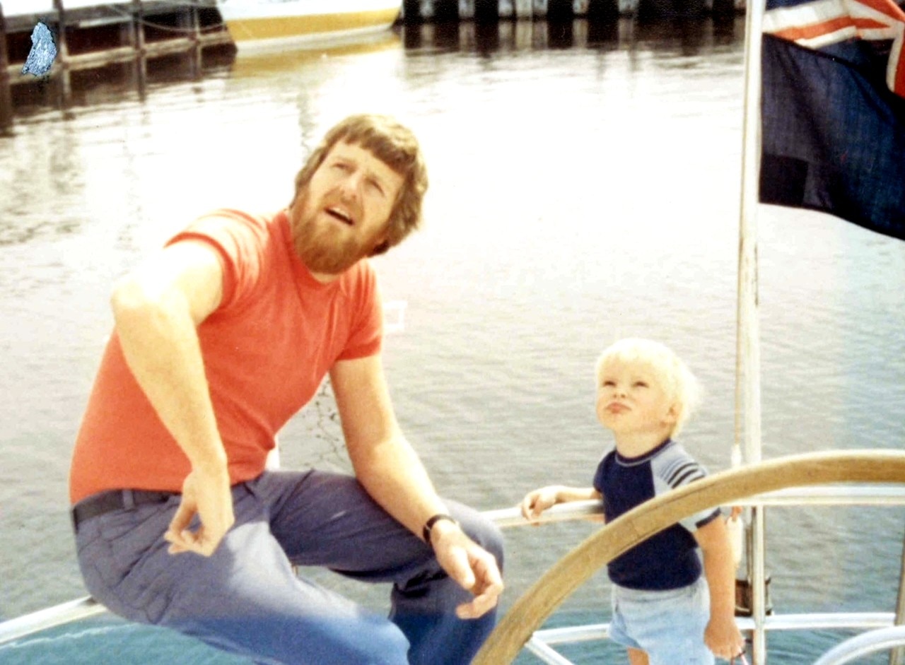 A man with a beard sits on a yacht next to a young blonde haired boy in the 1970s
