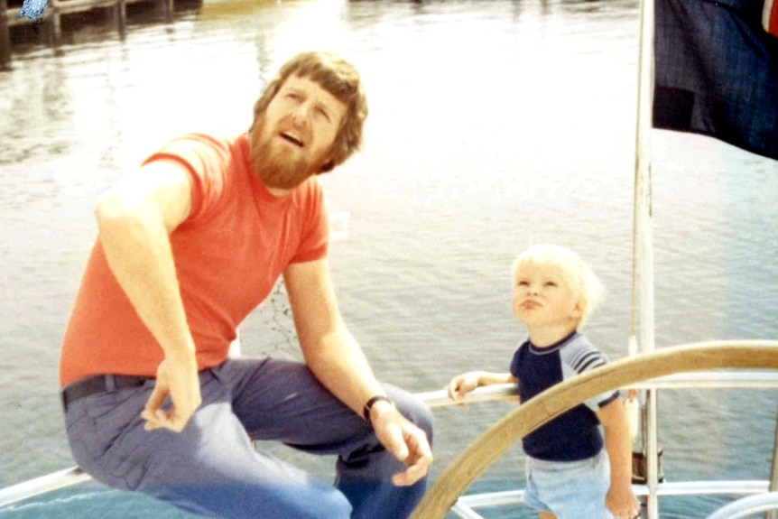 A man with a beard sits on a yacht next to a young blonde haired boy in the 1970s