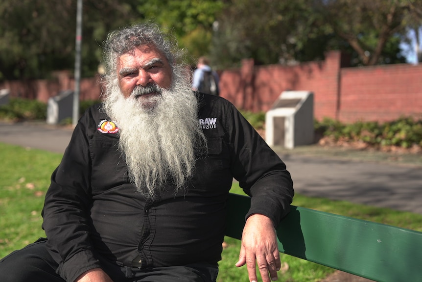 A man with a white beard in a black shirt sitting on a bench in front of a footpath