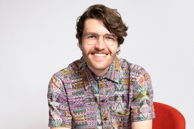 A smiling man, bespectacled, moustachioed and wearing a busy shirt, sits in a chair.