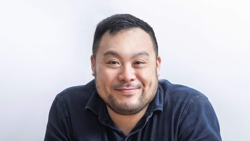 The chef David Chang wearing a blue shirt sitting at a bare table