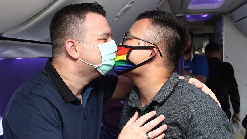 Two men wearing masks kiss on board an aeroplane