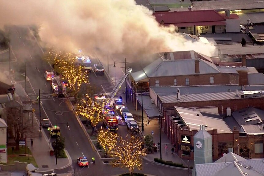 An aerial fire of smoke and fire coming out of a building surrounded by several fire trucks.