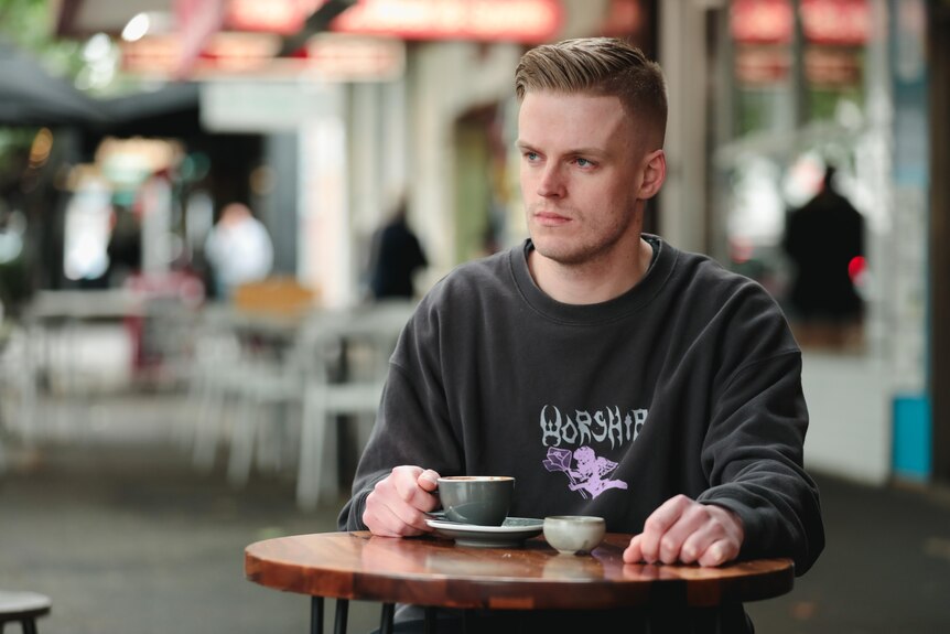 A young man sits a cafe table outside, his hand on a coffee cup. He looking to his right with a neutral expression.