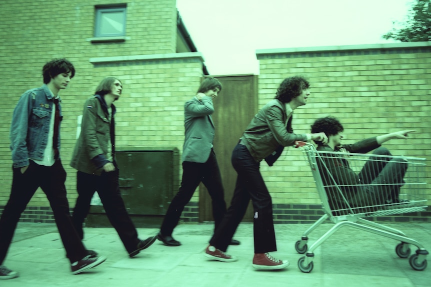A photograph of five young men, with a green wash over it. One is pushing another in a trolley, the other three walk behind.
