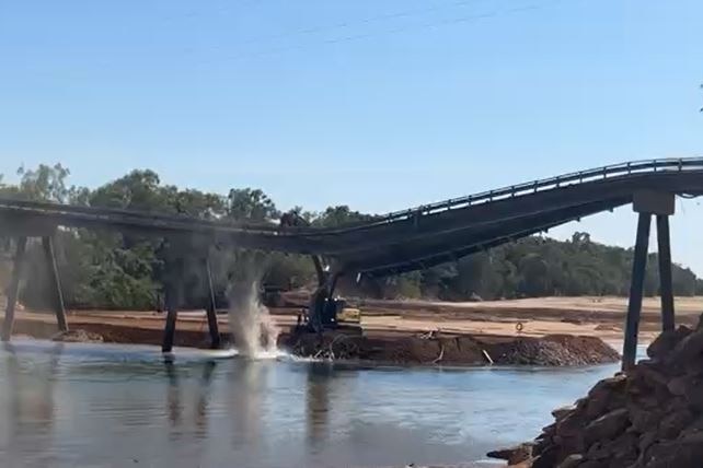 An excavator demolishing a bridge over a river.