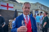 Nigel Farage stands holding a beer in a pub wearing a blue suit with a red waistcoat smiling at the camera