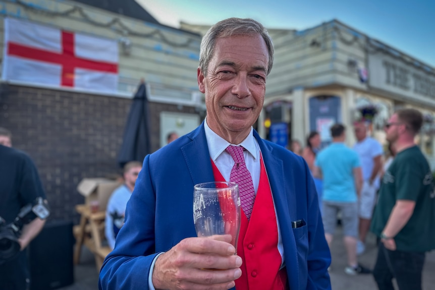 Nigel Farage stands holding a beer in a blue suit with a red waistcoat smiling at the camera