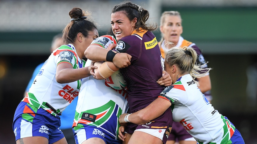 A Brisbane Broncos NRLW player stands with the ball as she is tackled by three Warriors opponents.