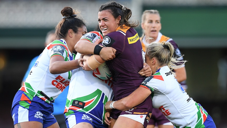 A Brisbane Broncos NRLw player stands with the ball as she is tackled by three Warriors opponents.