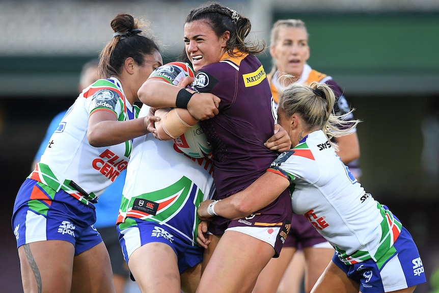 A Brisbane Broncos NRLw player stands with the ball as she is tackled by three Warriors opponents.