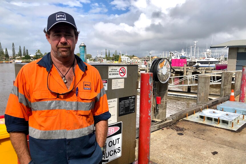 A man in a cap and a high vis shirt stands in front of a fuel bowser with no customers at the dock.
