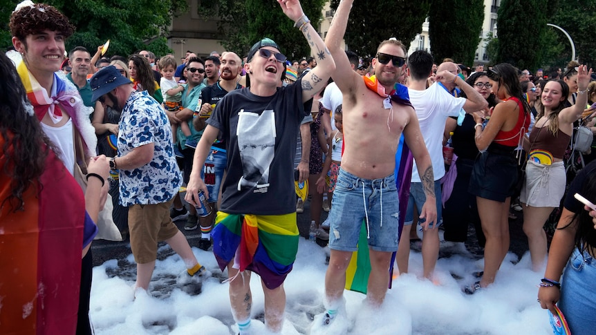 two men with sunglasses on wearing pride flags stand in bubbles on a street surrounded by a crowd