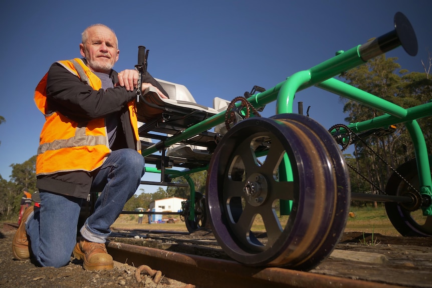 A man in a high-vis vest crouched on a rail track, leaning on a bright green rail bug.