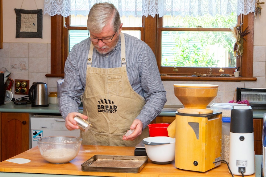 A greying man works around bowls and dough boards on a kitchen bench.