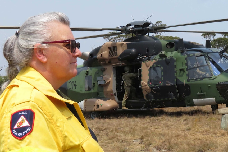 A woman in hi-vis yellow clothing stands in front of a military helicopter.