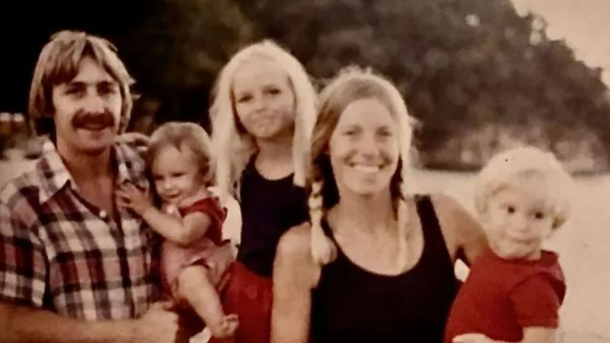 A mother and father smile at the camera with their three small children. Nature is behind them. 