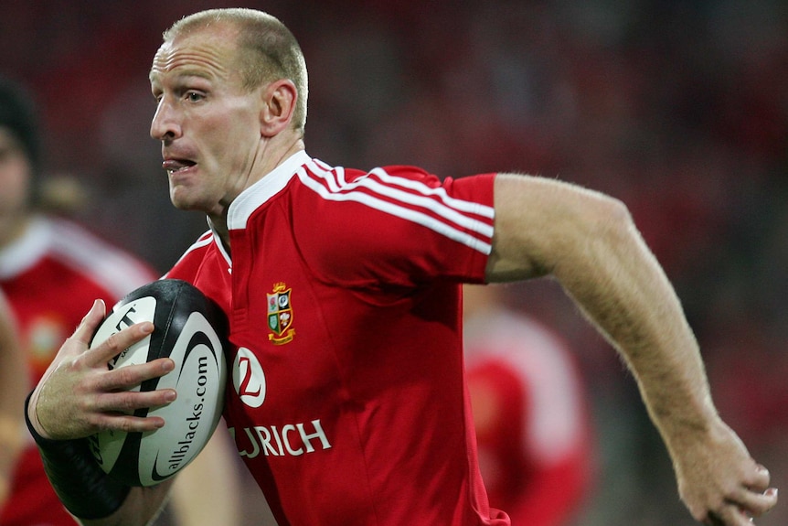 A British and Irish Lions player holds the ball with is right arm while playing against the All Blacks.