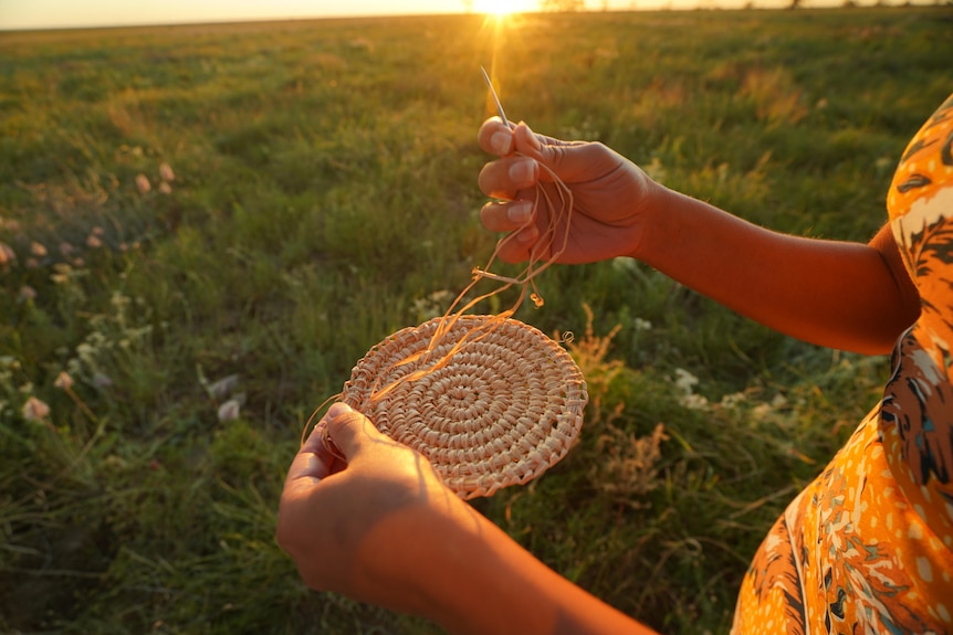 Woman weaving a basket in a field at sunset