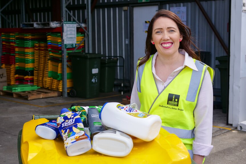 A smiling woman wearing a fluro yellow Brisbane City Council vest while standing next to a recycling bin.