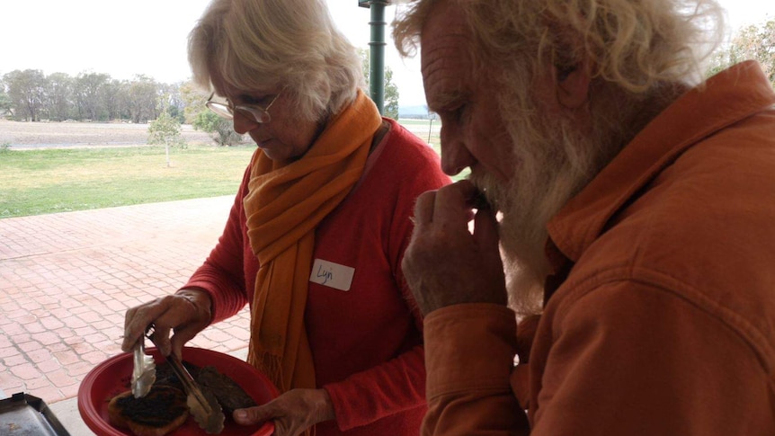 An older man and woman dish up food at an outdoor table as the man tastes something with his hands.