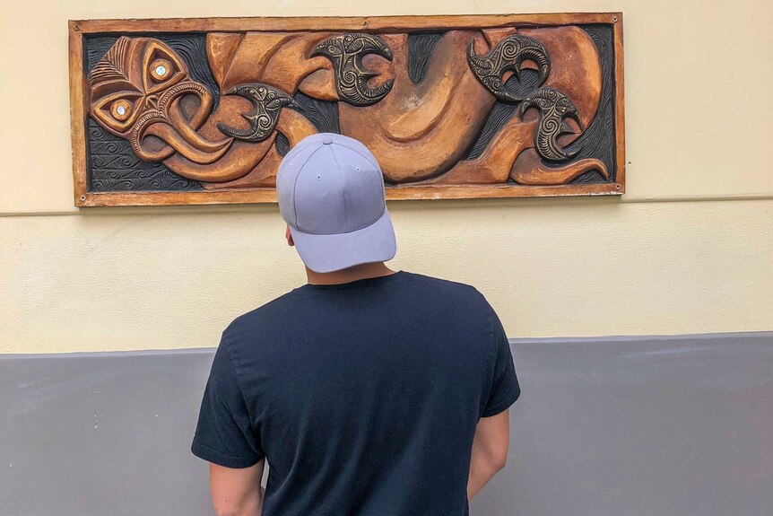 A young man wearing a cap backwards looking at a wooden carving hanging on the wall.