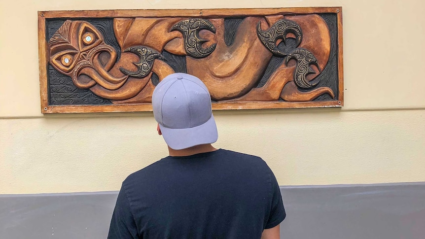 A young man wearing a cap backwards looking at a wooden carving hanging on the wall.