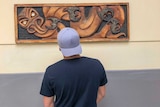 A young man wearing a cap backwards looking at a wooden carving hanging on the wall.