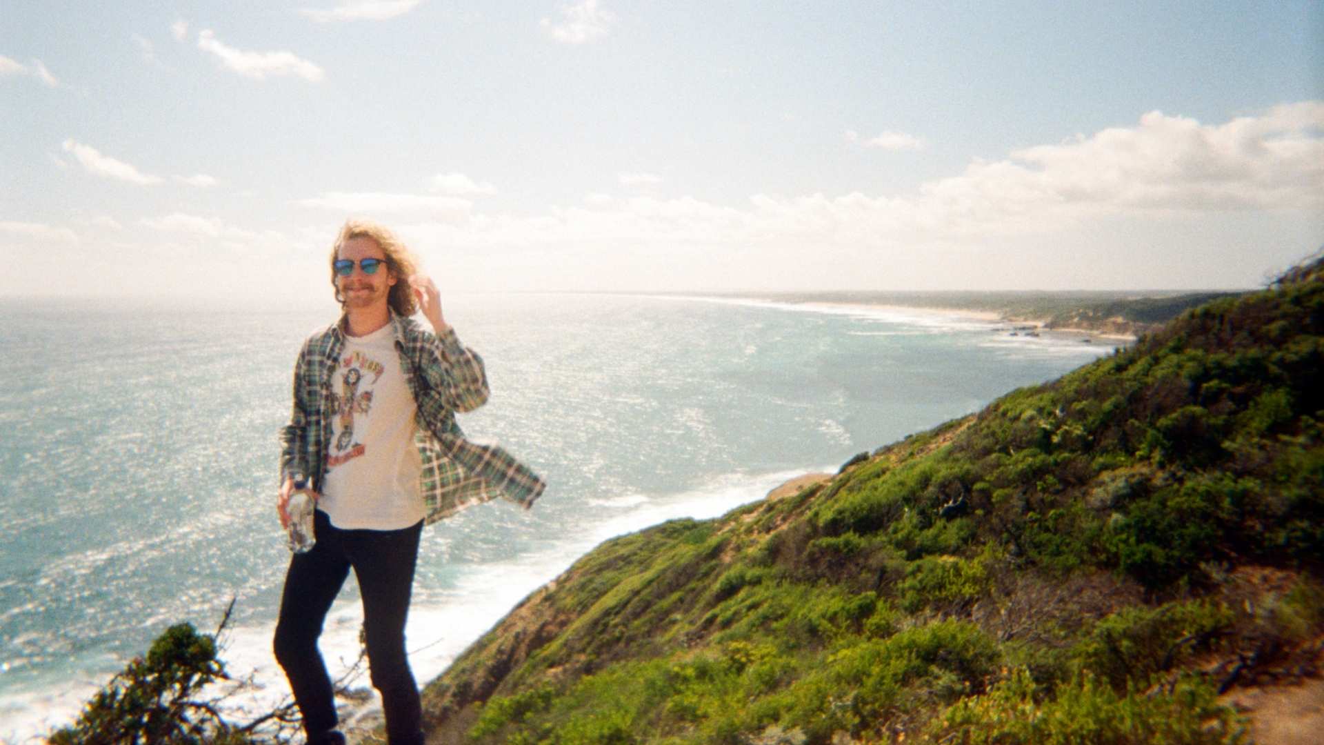 A young man with long curly hair and sunglasses stands on the edge of a cliff